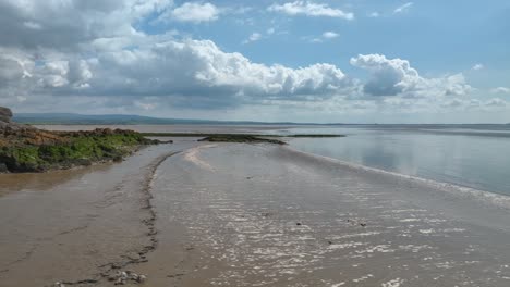 Seitlicher-Tiefflug-Entlang-Schlammigem-Strand-Mit-Flauschigen-Wolken-Am-Horizont-An-Einem-Hellen-Frühlingstag-Bei-Jenny-Brown&#39;s-Point,-Silverdale,-Lancashire,-England,-Großbritannien