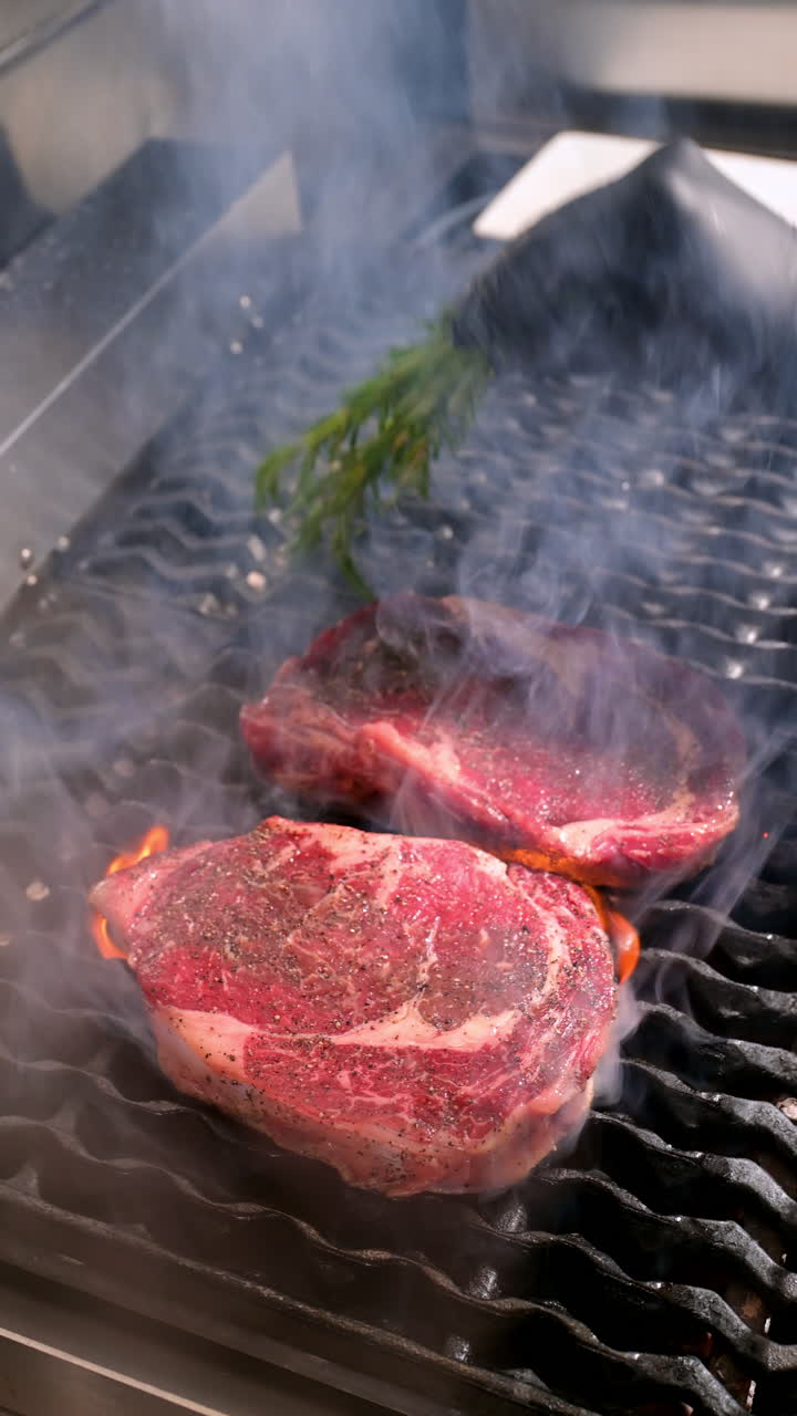 Raw ribeye steaks on grill with herbs. Two raw ribeye steaks with seasoning and rosemary placed on a smoky grill