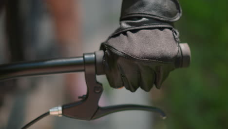 close-up of individual gripping bike handlebar with gloved hand, throttling and holding brake lever, with sunlight subtly reflecting off the glove, background features a soft blur of greenery