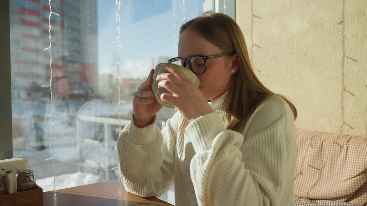 Side view of professional woman in white sweater holding coffee cup while seated in modern cafe during morning, sunlight casting shadows, with cityscape and snowy background