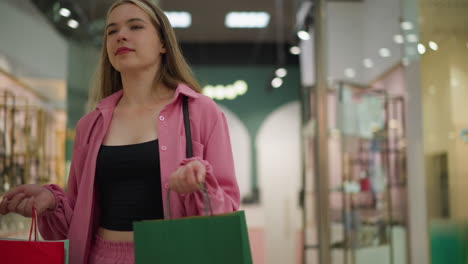 lady in pink dress and shorts holding red, green, and mint green shopping bags with a warm smile, walking through a brightly lit mall with reflections of lights on glass in the background