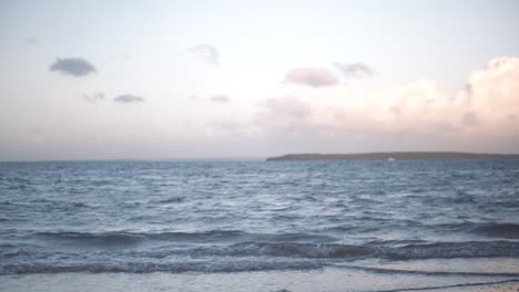 water gently sloshing itself onto the beach at sunet as the retreating sun spills pastel colours of pink and blue into the sky and fluffy clouds whilst an island and boat sit off in the distance