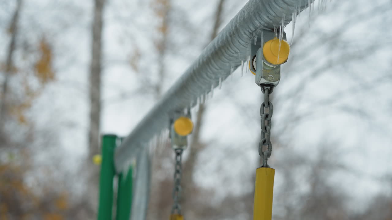얼음과 얼음으로 인 노란색 막이있는 금속 스 체인 관절의 클로즈업은 얼어붙은 금속 바를 따라 매달려 있으며, 벌거벗은 나무와 평온한 분위기와 함께 흐릿한 겨울 배경에 설정되어 있습니다.