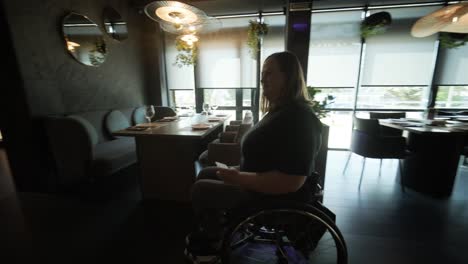 una mujer en silla de ruedas está sentada sola en un restaurante moderno, mirando por grandes ventanas. la imagen destaca la accesibilidad e inclusión en un elegante entorno de comedor con luz natural.