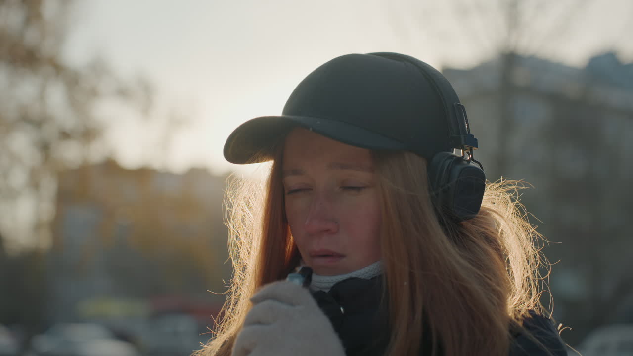 Red haired caucasian woman with headphones standing on urban sidewalk at dusk in winter coat and gloves. Backlit by low sun, breath visible in cold air, adjusting scarf while listening to music