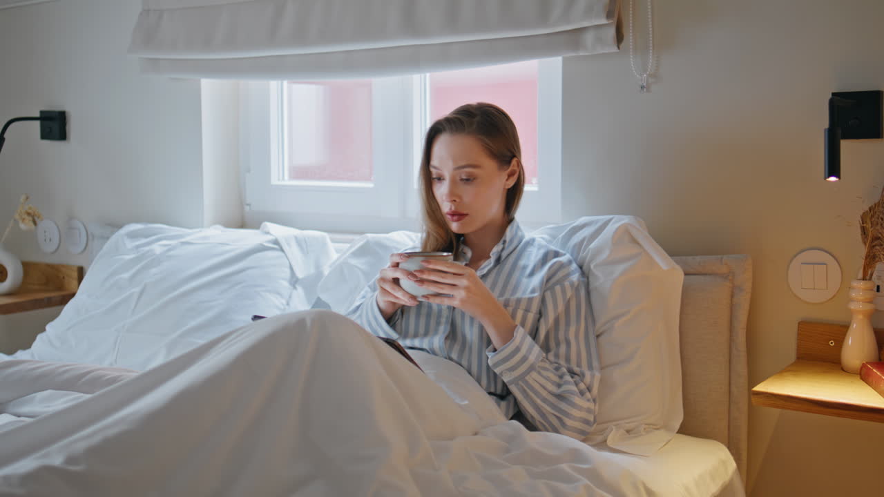 Lazy girl drinking coffee in cozy bed at morning. Calm woman reading tablet