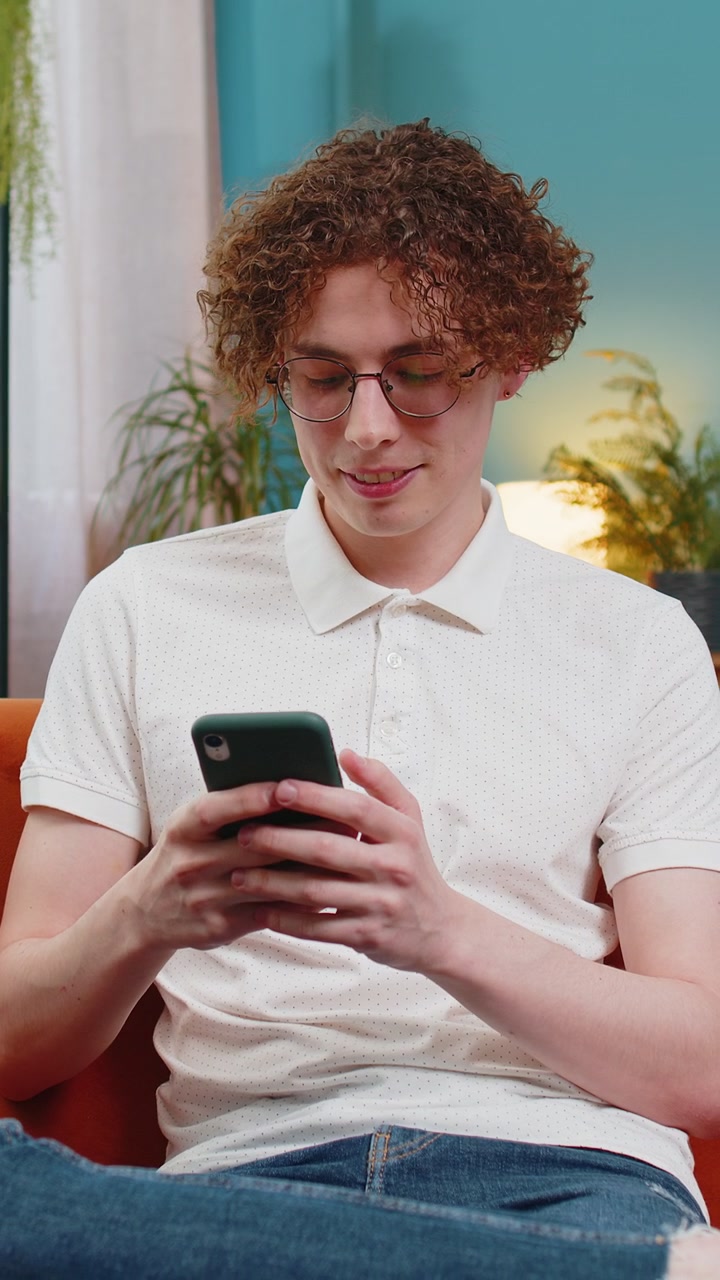 Giovane caucasico seduto sul divano usando lo smartphone mobile condivide messaggi sorride nella stanza di casa