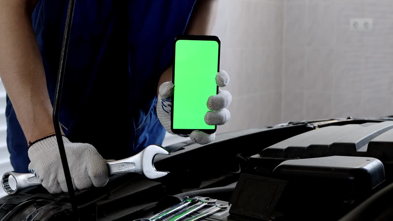 un mecánico de automóviles con uniforme y guantes sostiene un teléfono inteligente con una pantalla verde hacia adelante. reparación de automóviles en el garaje usando equipos. problema de motor abierto del capó.