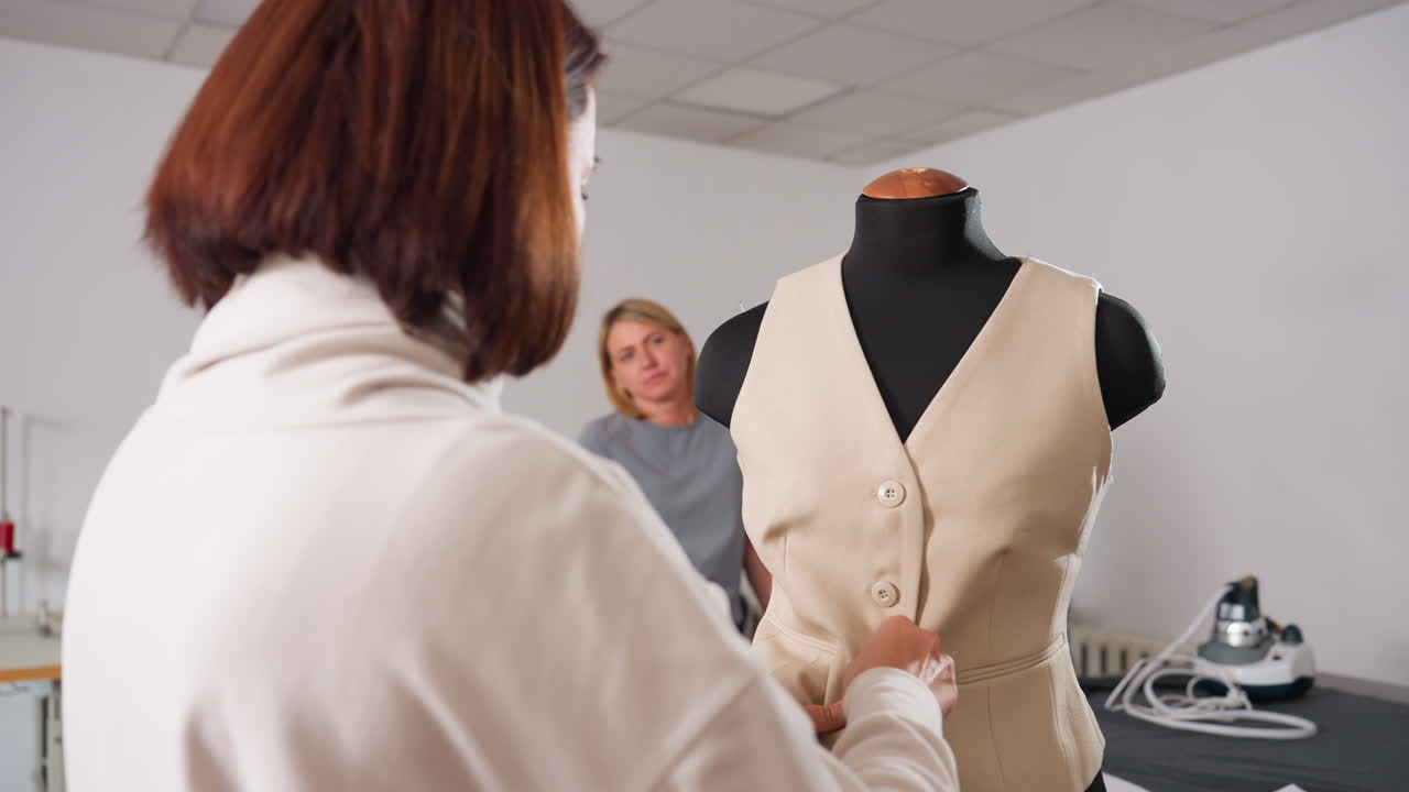 Two cloth stylists in fashion studio as one admires tailored beige jacket on black dress form while other unbuttons and prepares to remove garment, demonstrating craftsmanship and teamwork