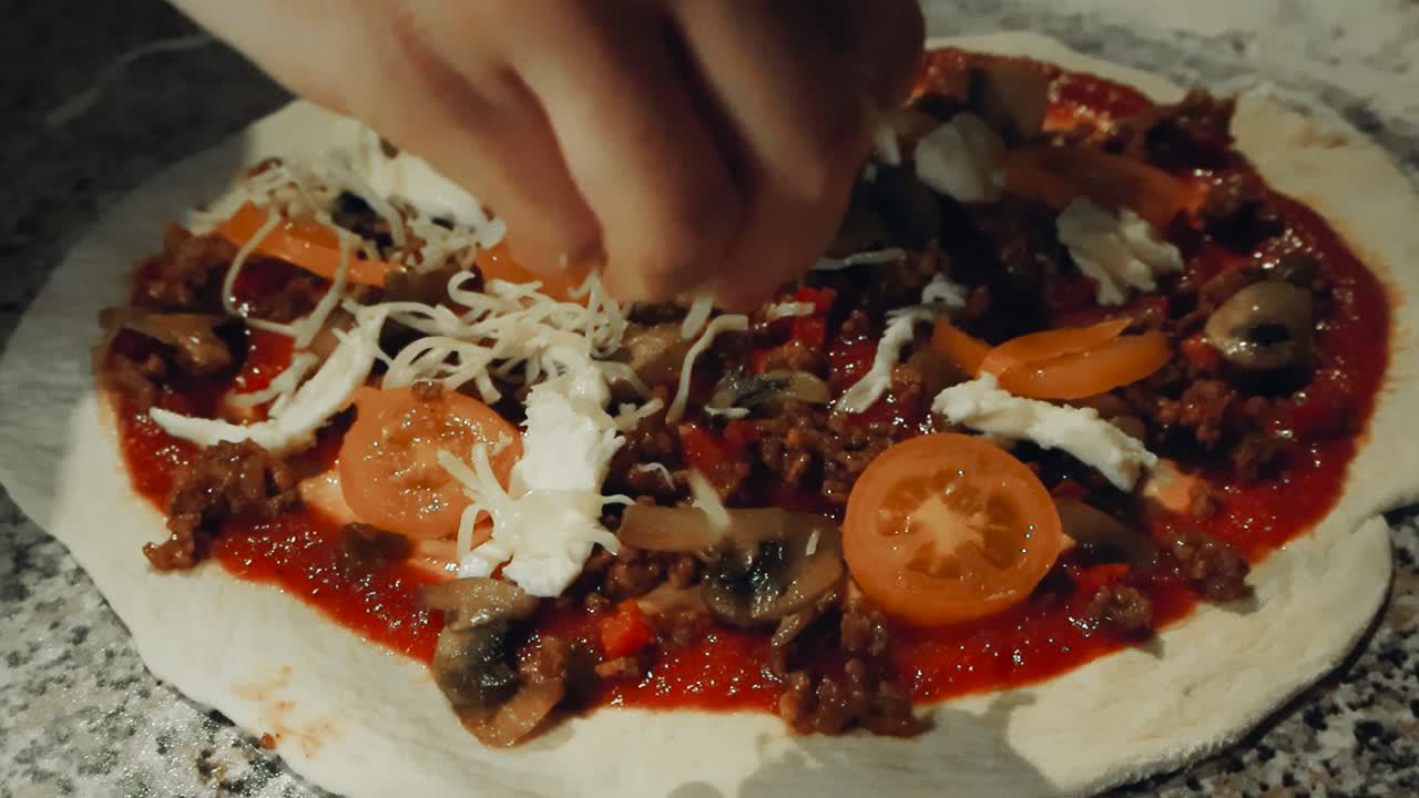 Close up video of a chef or home cook hand spreading delicious cheese and mozzarella on a home made pizza on a countertop in slow motion that has white flour around. Tomatoes, meat and sauce visible