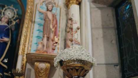 white paschal candle adorned with flowers stands before gold religious art in church