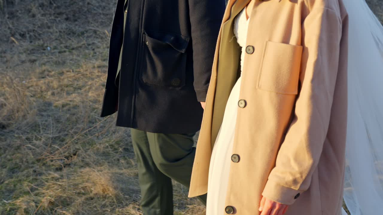 Close up couple holding hands walking on dry grass in warm coats with hidden wedding dress and suit underneath in sunny outdoor romantic atmosphere