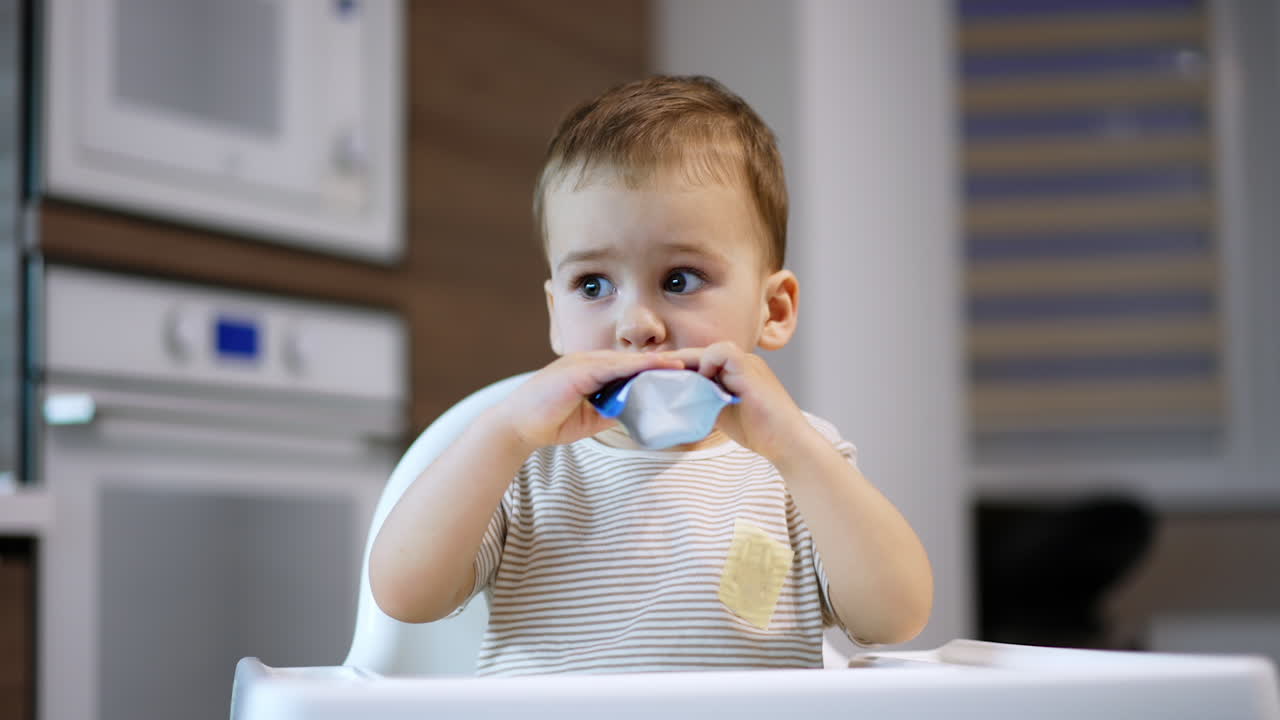 Adorable baby boy takes a pack of food into mouth. Little kid eating the pureed fruit willingly. Blurred backdrop.