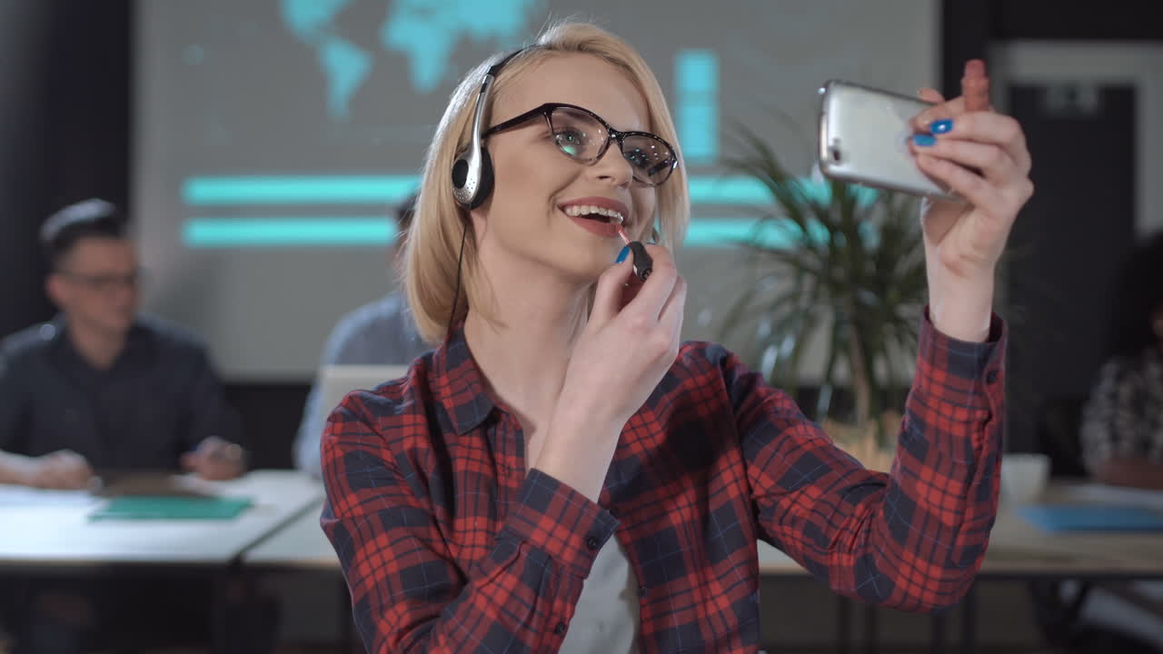 Woman applies makeup using her smartphone as a mirror in an office