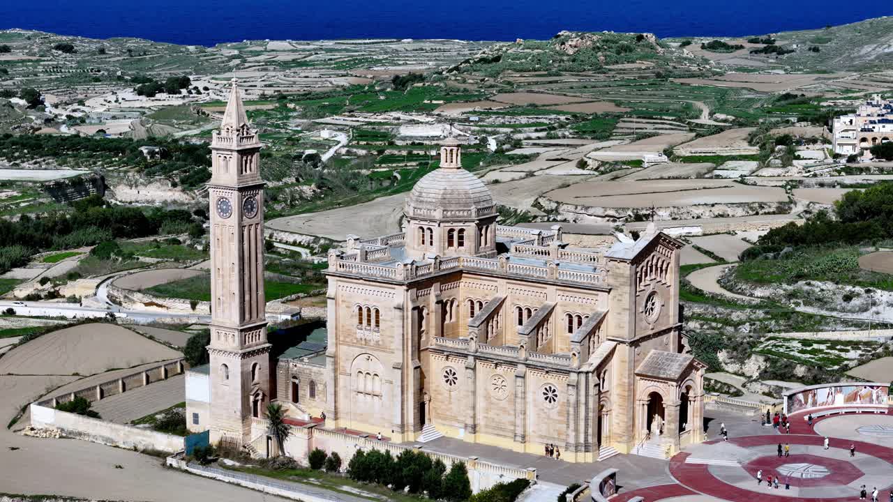Cinematic wide high angle shot of the majestic Ta Pinu Basilica in Gozo Malta during a golden summer afternoon under a clear blue sky featuring warm limestone textures and lush green terraced fields