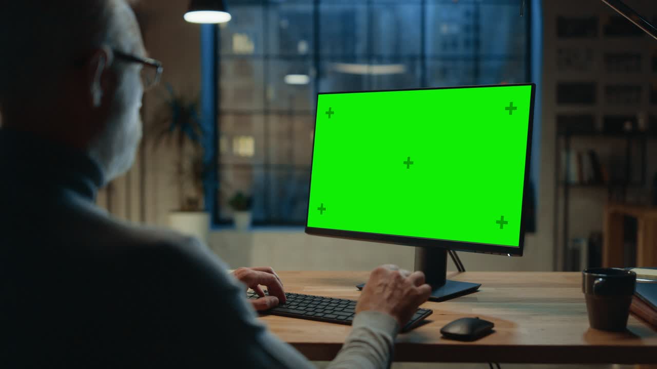Over the Shoulder: Confident Middle Aged Man Sitting at His Desk Using Desktop Computer with Mock-up Green Screen. Evening in the Stylish Office Studio with City Window View