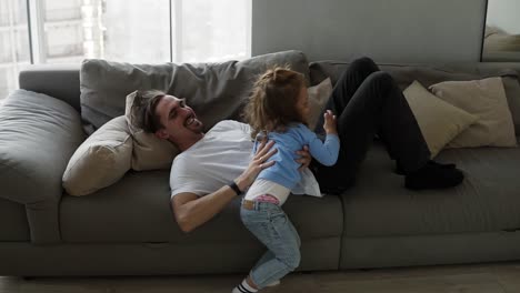 Happy-family-father-and-kid-daughter-having-fun-together-lying-on-sofa-father-tickles-the-girl,-raises-in-his-arms,-and-she