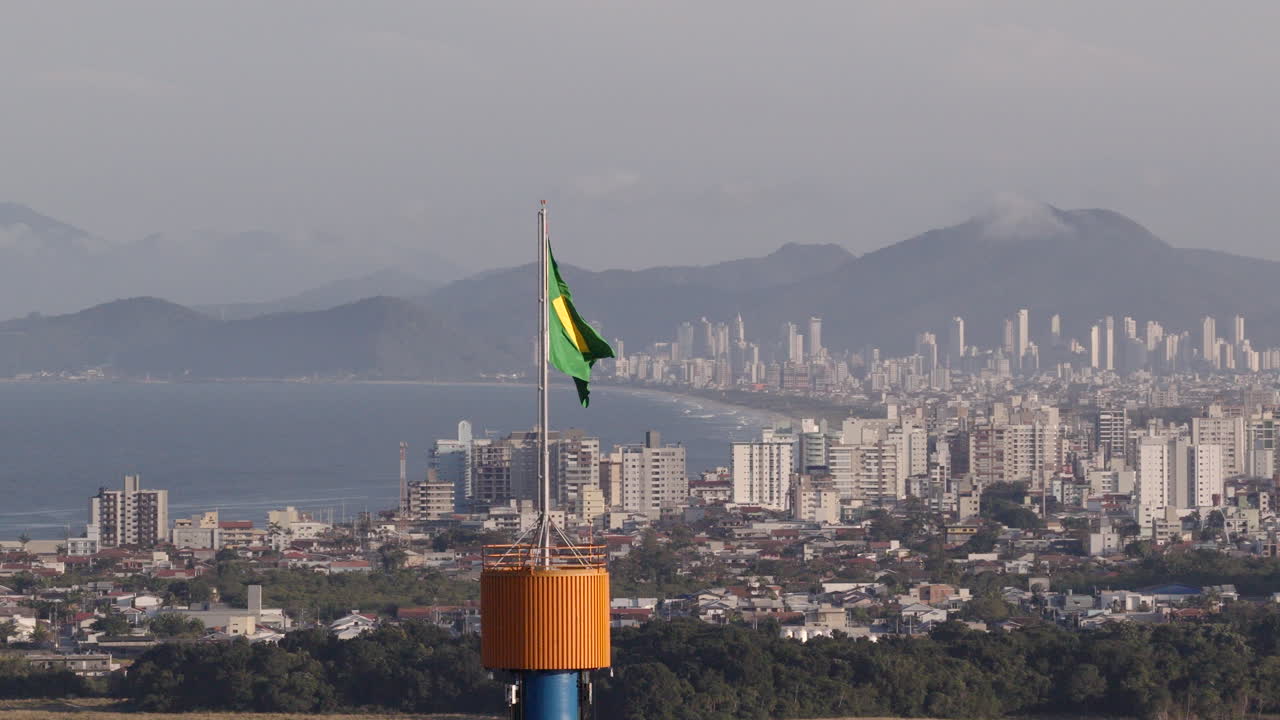 Brazilian flag waving above city skyline with ocean and mountains in background