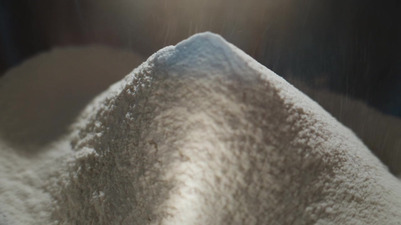 Wit poeder stroomt zachtjes op een hoop, vastgelegd in slow motion. Stof valt zachtjes, creëert textuur en zachte focus. Ideaal voor bakken, koken of industriële processen. Perfect voor voedsel, wetenschap.
