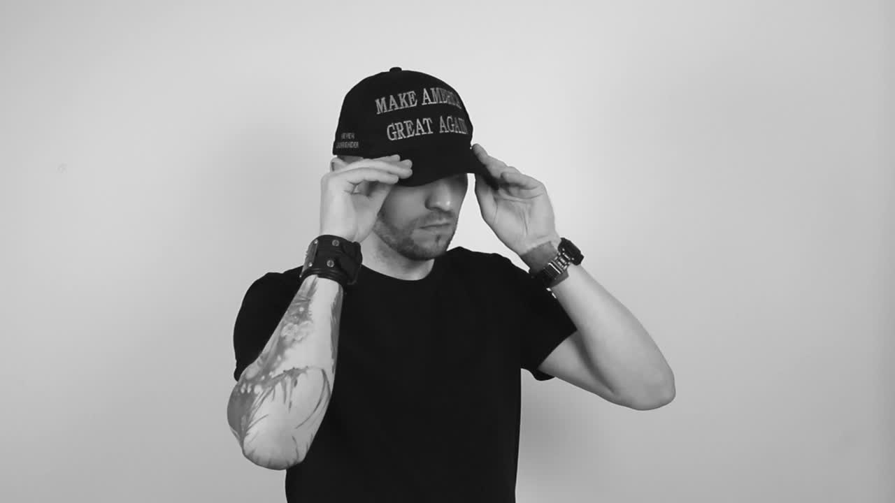 Grayscale footage of a tattooed man with short beard and hair putting on a dark black MAGA hat or hat with Make America Great Again writing on it, man crosses arms and nods with confidence, approval.
