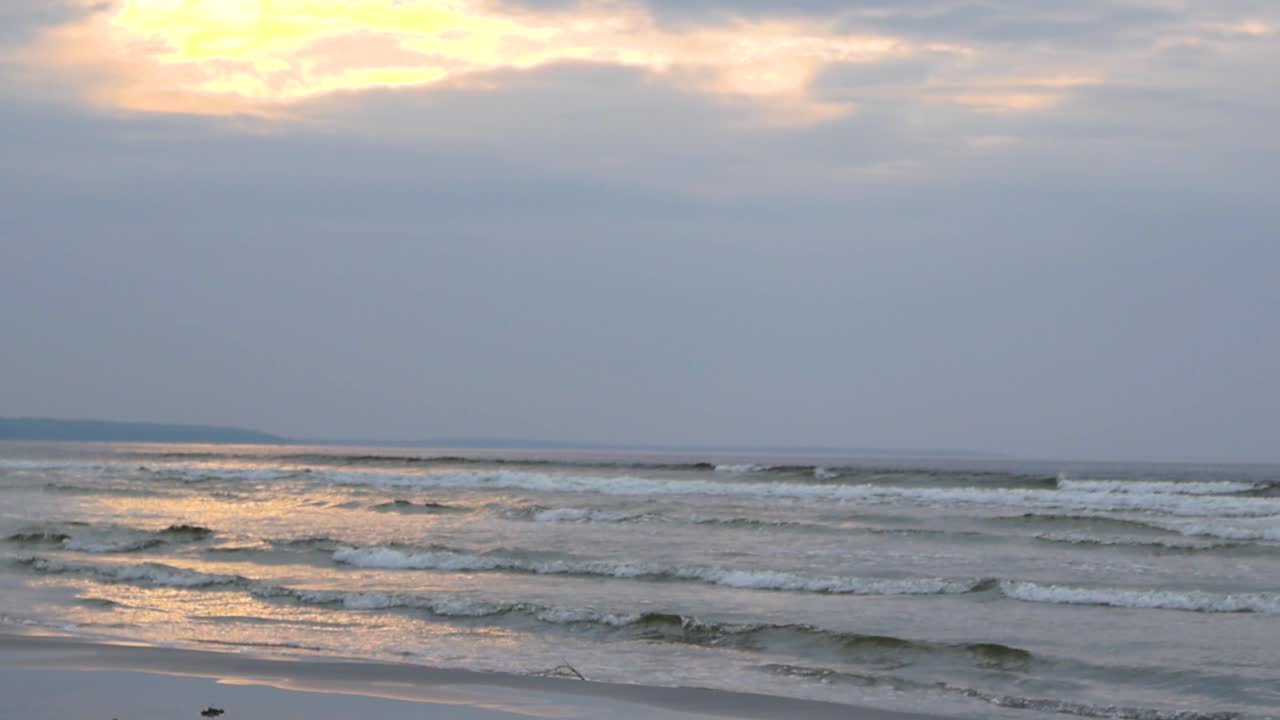 Gorgeous slow motion footage of large ocean sea water foamy and bubbly white water waves coming to a sandy beach shoreline during sunset while the sky is slightly orange colored and reflects on water.