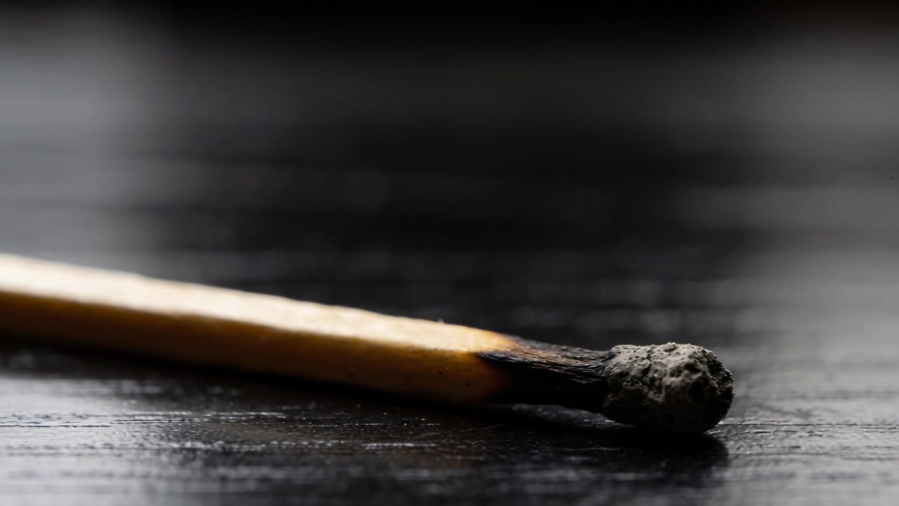 La messa a fuoco si sposta, la telecamera rivela un fiammifero di legno bruciato su un piano d'appoggio di legno scuro, mostrando la testa carbonizzata