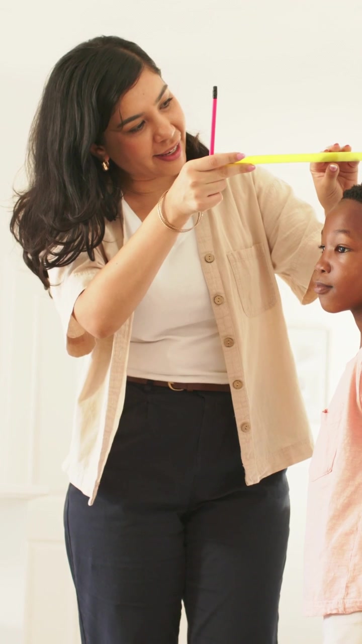 Vertical video: Placing yellow ruler, woman measuring boy in pink T-shirt, marking with pink pencil