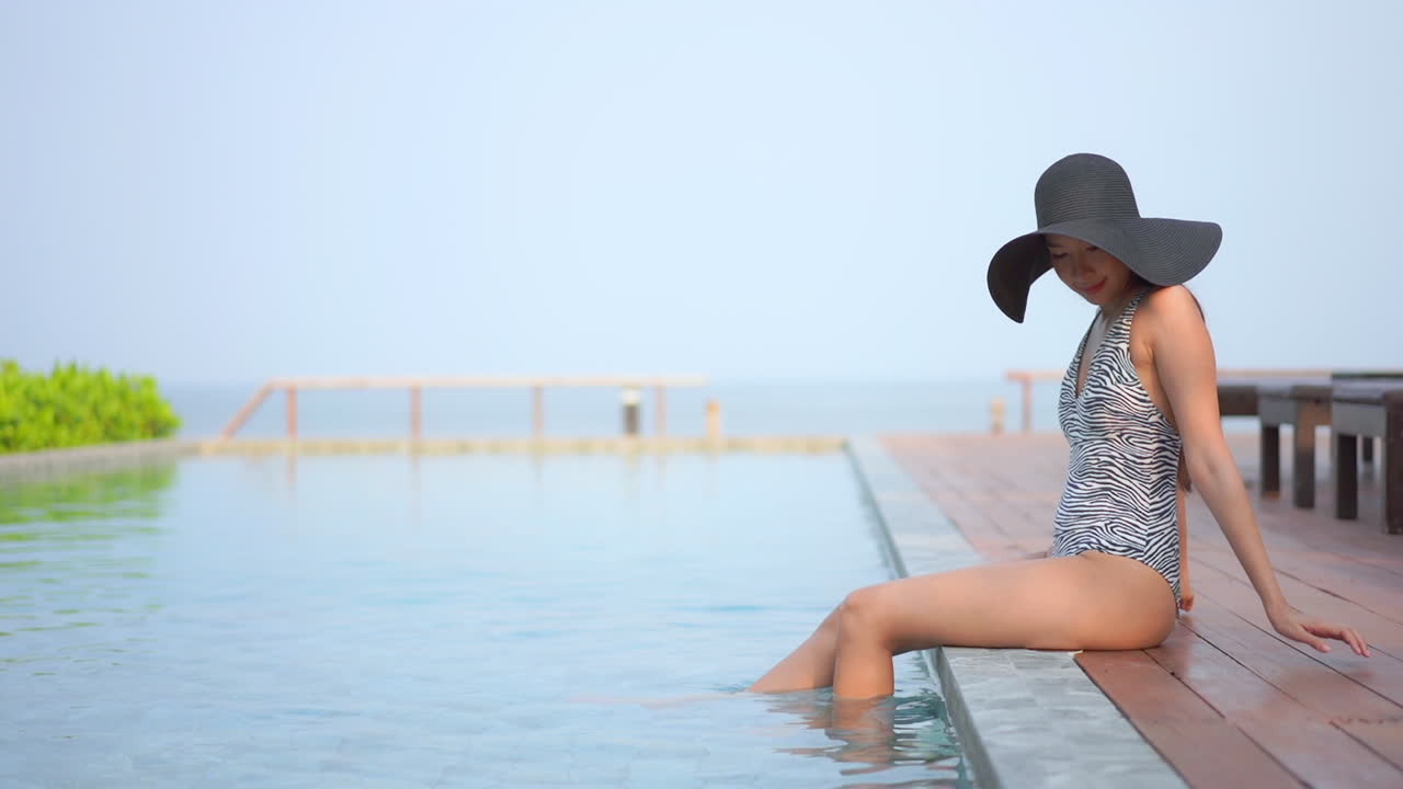 A young woman in a one-piece swimsuit and huge sun hat sits on the edge of a resort pool playfully kicking her feet in the water. Copy space