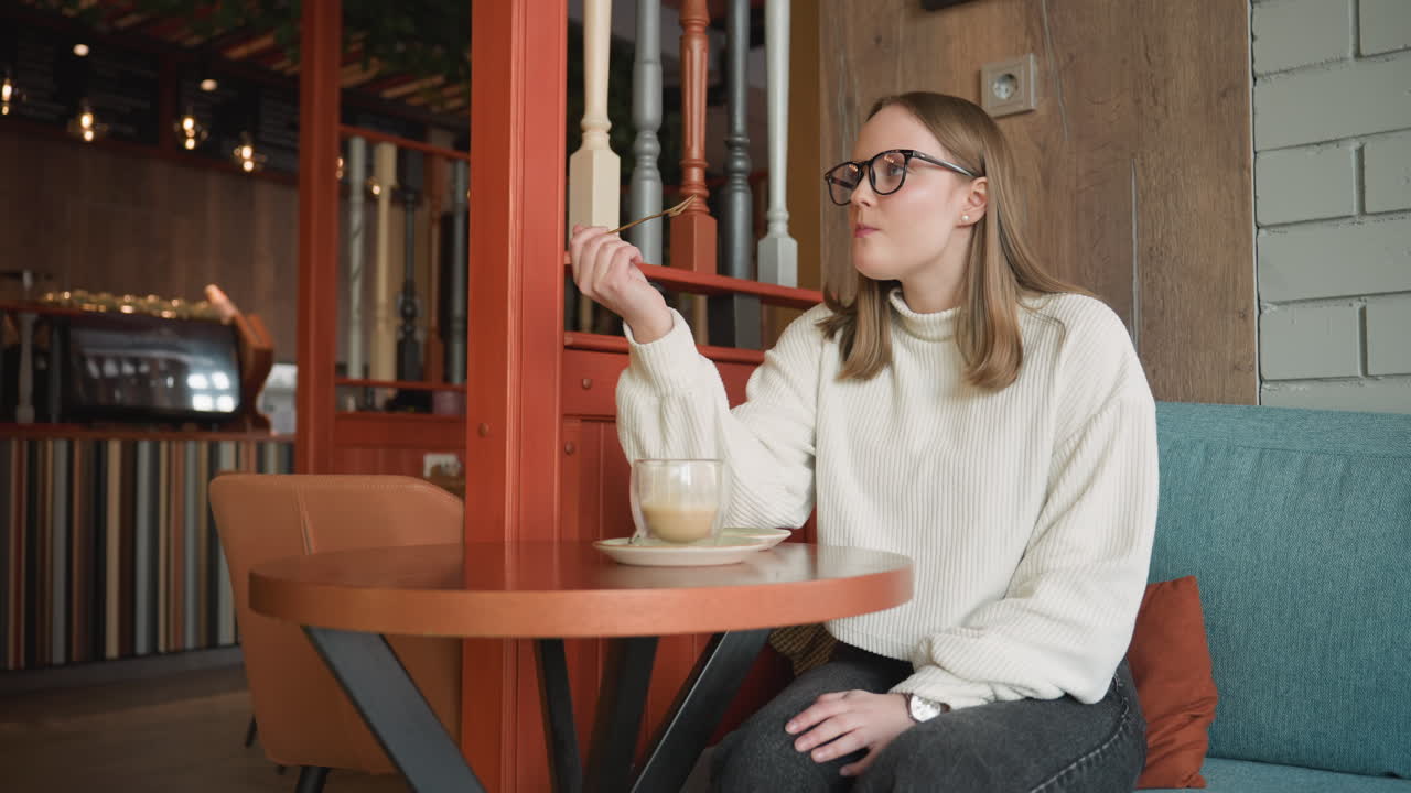 young woman in white sweater sits calmly in cozy cafe holding glass cup of latte, enjoying dessert while seated on teal sofa near red wooden partition and warm ambient lighting in stylish interior