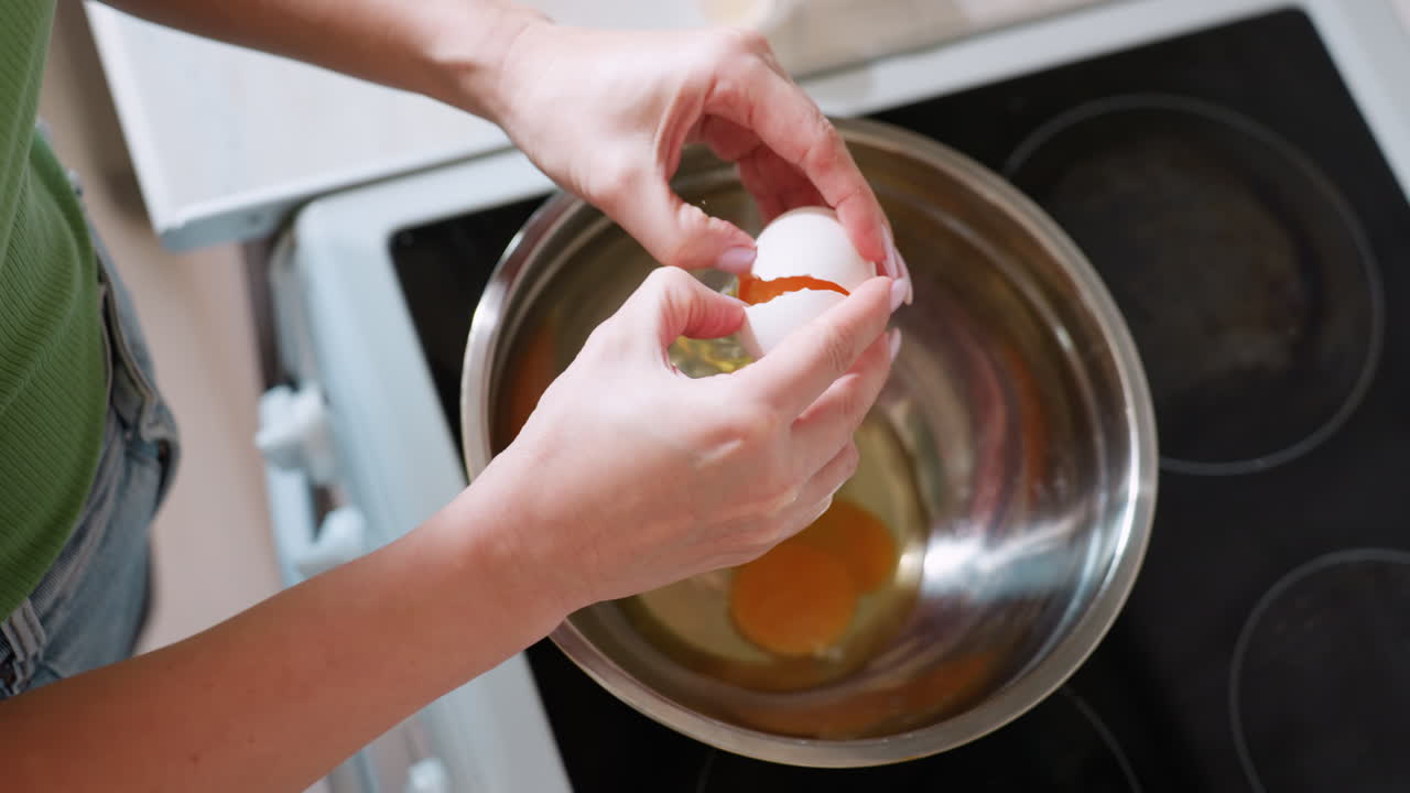Top down view of woman hand carefully opening raw egg over stainless steel bowl in kitchen, yolk dropping into container while preparing ingredients for cooking recipe with attention to detail