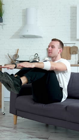 young man doing workout at home. handsome guy sitting on a couch and doing exercise during quarantine in the kitchen. vertical video