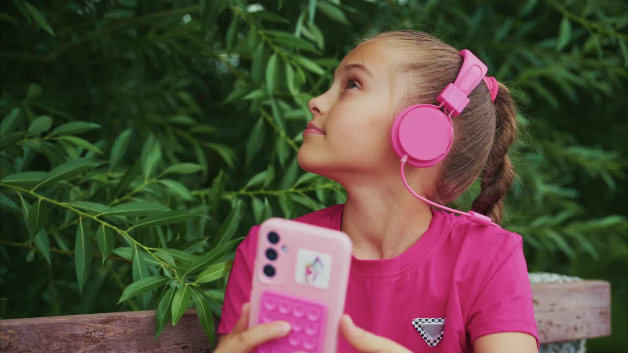 A Young Girl Enjoying Music with Pink Headphones and a Phone, Surrounded by Lush Greenery in a Relaxing Outdoor Setting