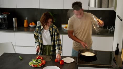 A-happy-young-man-with-Black-skin-in-a-cream-T-shirt-together-with-his-girlfriend-an-adult-young-brunette-is-preparing-breakfast.-The-man-opens-the-lid-of-the-frying-pan-and-thick-steam-comes-out-from-there-in-the-kitchen