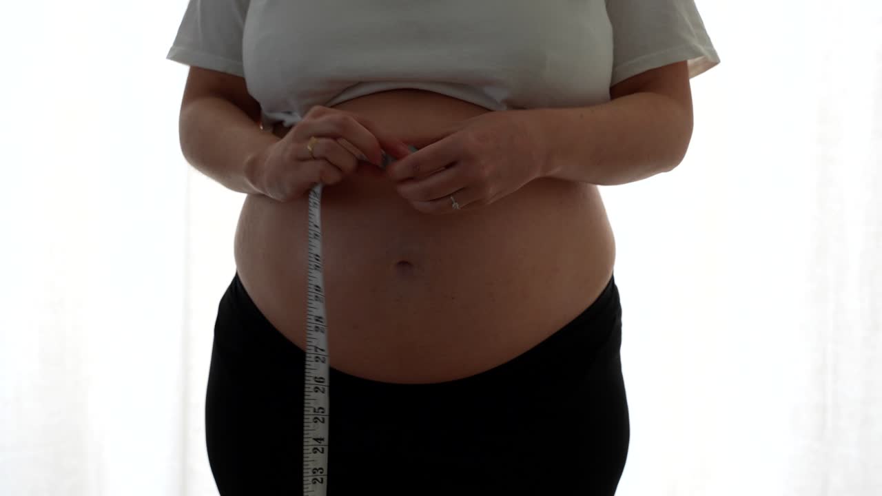 Pregnant woman rolls tape after measuring her belly at 38 weeks in soft daylight