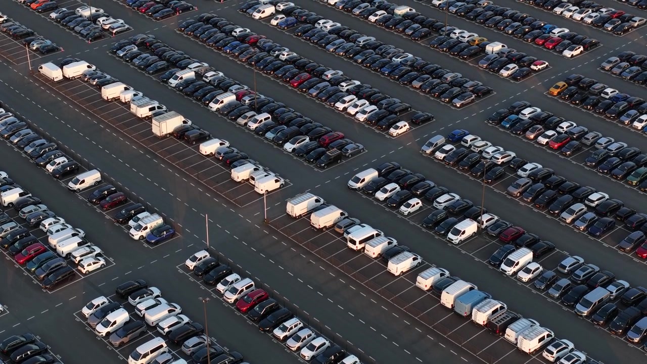 Fugleperspektiv der viser hundredvis af splinternye biler og varevogne perfekt opstillet på en enorm parkeringsplads ved en bilfabrik, der venter på forsendelse og distribution til forhandlere.