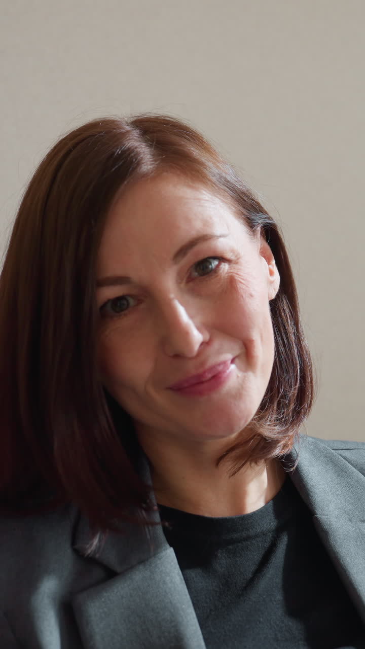 Businesswoman in gray blazer sitting on couch using tablet in quiet modern office setting, focused on screen with natural light casting soft shadows across face, indoor plant partially visible
