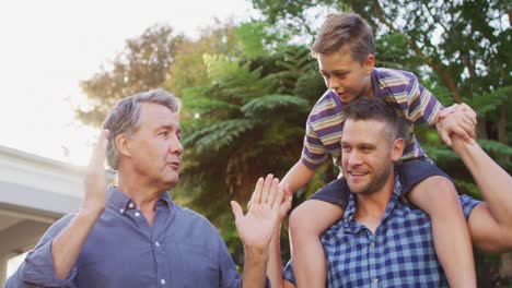 happy caucasian grandfather talking to grandson, sitting on father's shoulders in garden