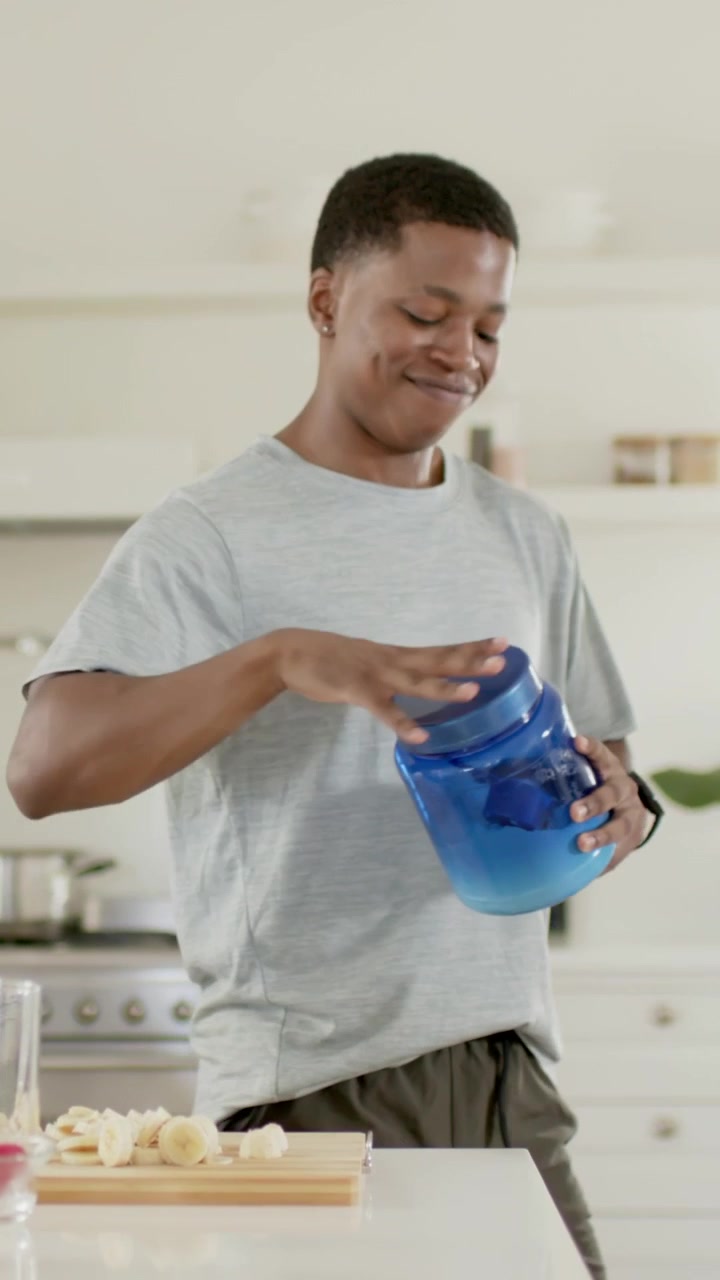 Video vertical: Hombre con un recipiente azul de tapa giratoria llevándolo a un armario superior, guardándolo después de un tentempié de plátano.