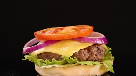 a sesame seed bun drops onto a beef cheeseburger with lettuce, tomato, onion, and cheddar, captured in slow motion against a black background with studio lighting