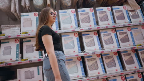 mujer joven caminando por el pasillo de la tienda, pasando por un estante lleno de productos de cama, mira a su alrededor, observando diferentes opciones de edredones y almohadas