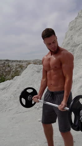 muscular shirtless man exercising hard in the mountains. bodybuilder with barbell on white mountain background in summer outdoors. vertical video