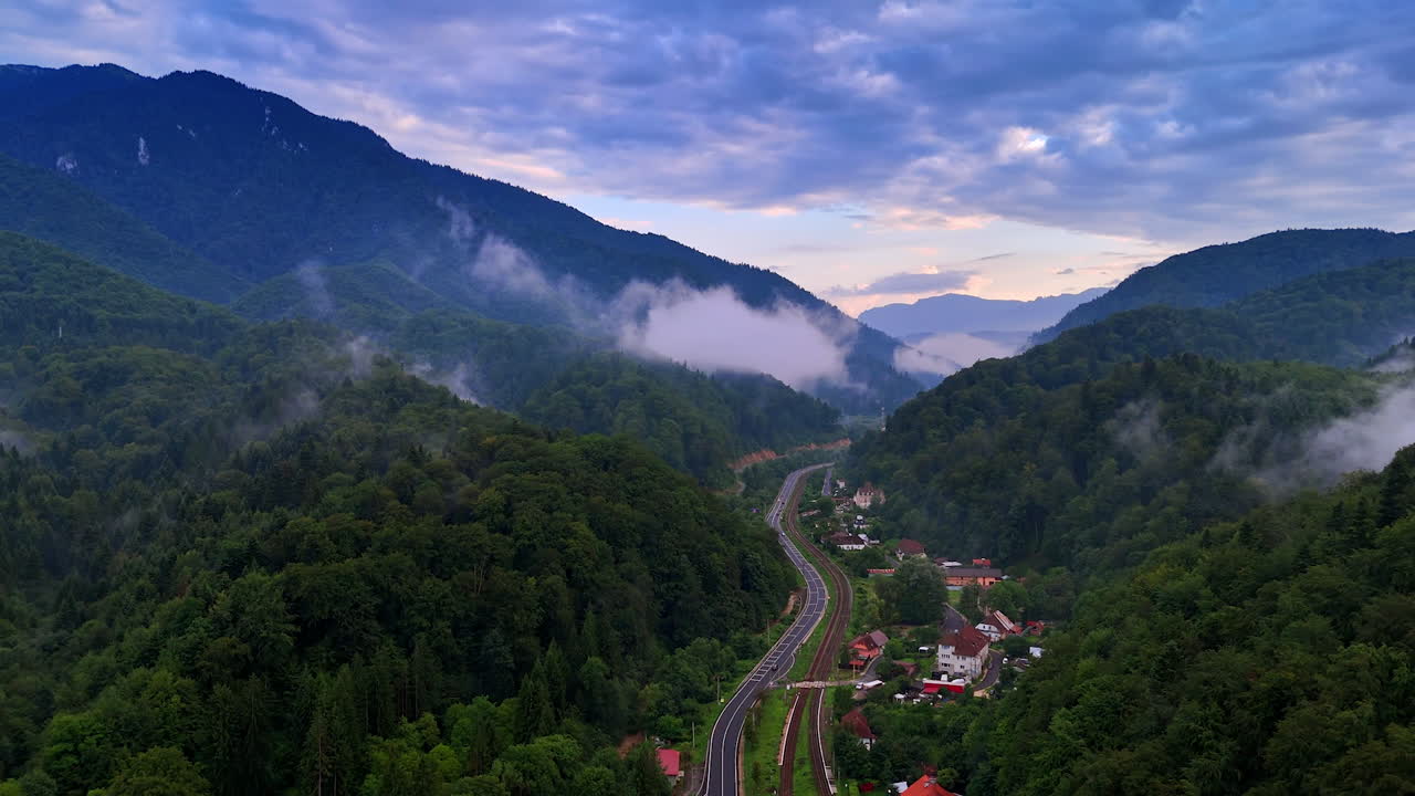 Footage over the residential area located among the wooded mountains. Drone flight over the railway and highway crossing the village. Dramatic cloudscape covers the sky over the scenery. Romania countryside