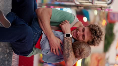 Vertical-video-of-a-man-with-curly-hair-and-stubble-in-a-Green-T-shirt-hugging-his-little-blond-son-while-sitting-on-an-attraction-in-an-amusement-park