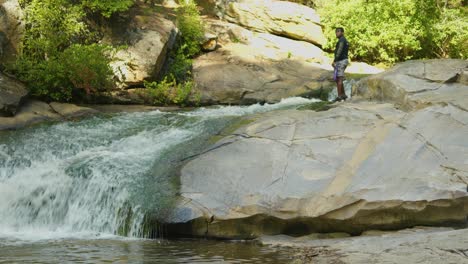 a man searches for something alongside a fast moving river in the middle of a lush forest