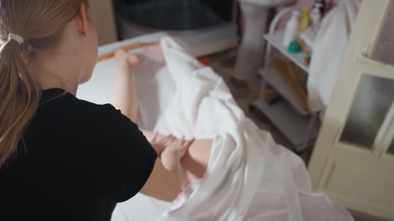 Overhead view of female massage therapist with tied-back blonde hair wearing black top gently massaging client thigh under white sheet in bright wellness room with skincare shelf