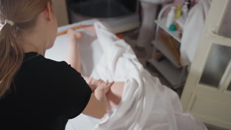 overhead view of female massage therapist with tied-back blonde hair wearing black top gently massaging client thigh under white sheet in bright wellness room with skincare shelf