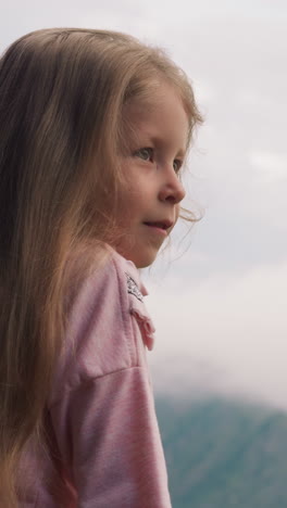 happy blonde little girl in pink sweatshirt admires low dense clouds covering mountains surrounding hotel at highland on overcast day close view slow motion