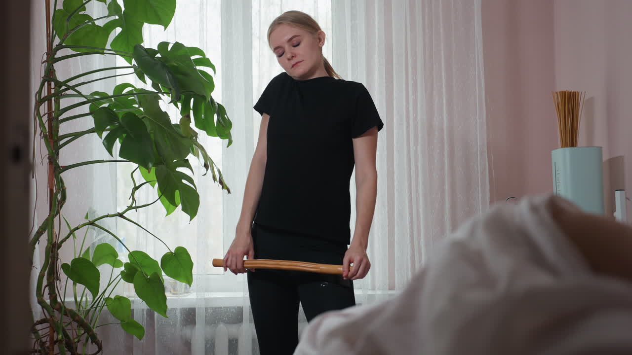 Massage therapist in black stretches neck for comfort while standing beside large indoor plant and holding massage stick in softly lit room, expressing calm body awareness and physical relief