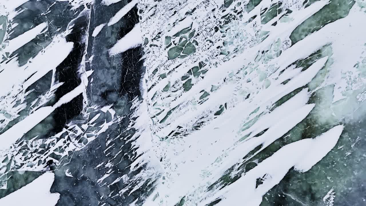 Primo piano drone della superficie marina ghiacciata con striature di neve e crepe scure, che creano una trama invernale astratta e un motivo naturale drammatico
