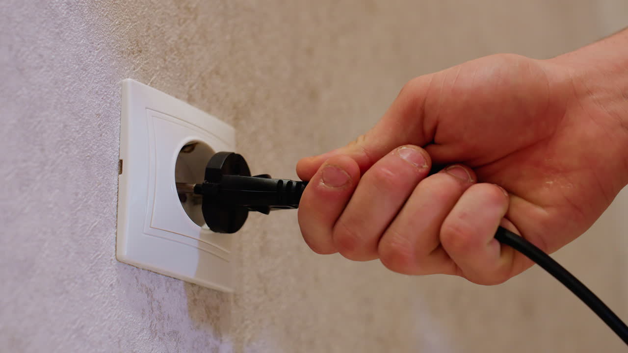 Close up of hand holding black plug wire inserting into wall socket on textured surface background, showing detail of electrical connection and household power usage during setup of appliance